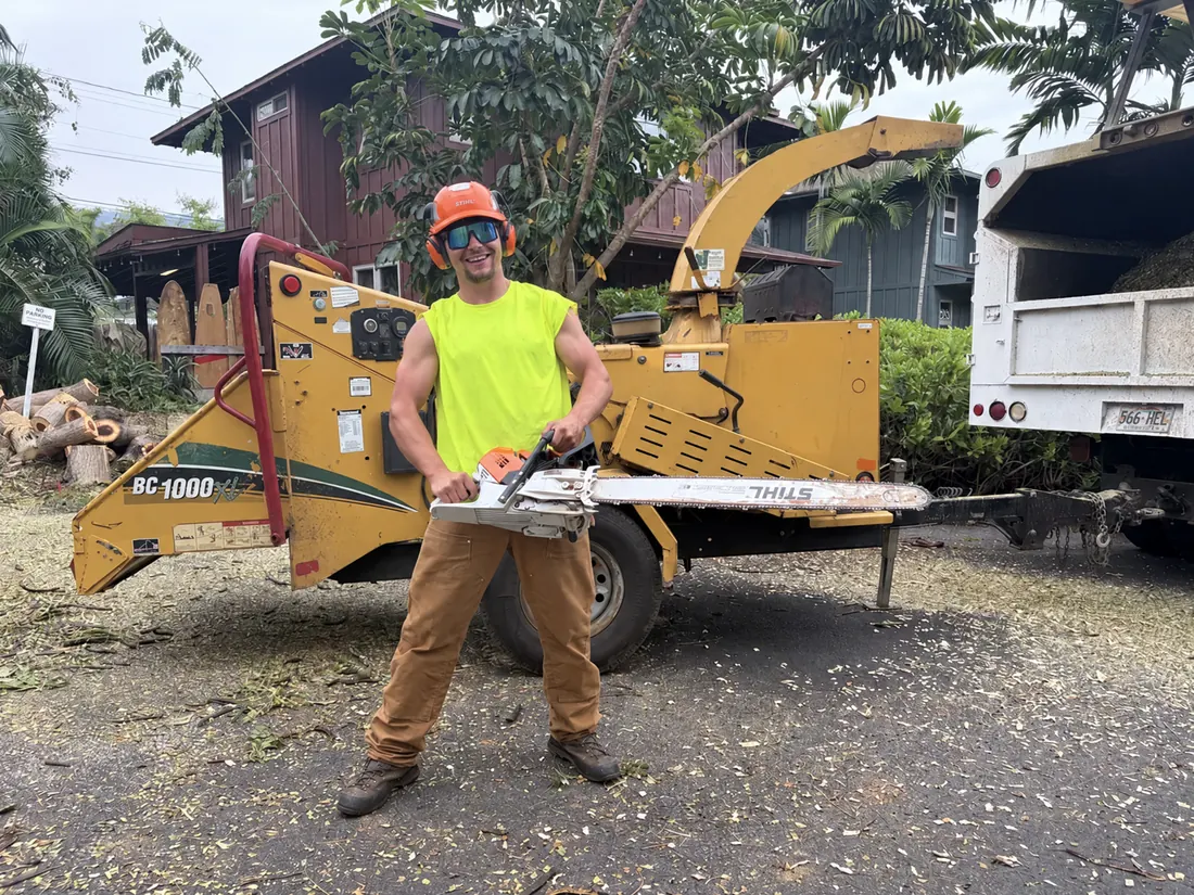 Worker holding a chainsaw beside a wood chipper and white dump truck